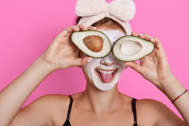 Молодая женщина держит две половинки авокадо и закрывает ими глаза, показывает язык, носит повязку для волос, имеет увлажняющую маску на лице, веселится, делая косметические процедуры дома.