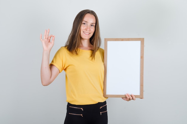 Young woman holding white board with ok sign in t-shirt, pants and looking glad.