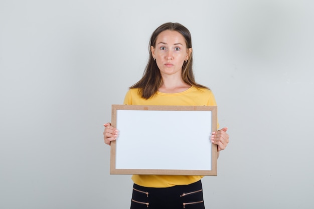 Young woman holding white board in yellow t-shirt, black pants and looking careful.