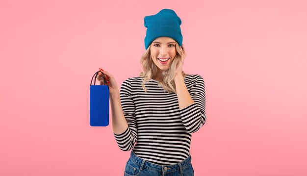 Young woman holding wireless speaker listening to music wearing striped shirt and blue hat smiling posing on pink