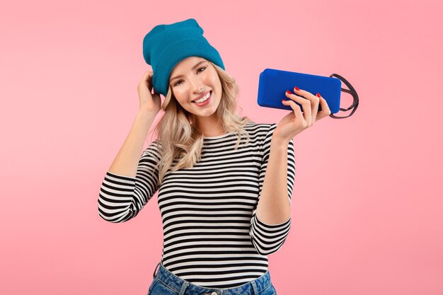 Young woman holding wireless speaker listening to music wearing striped shirt and blue hat smiling posing on pink