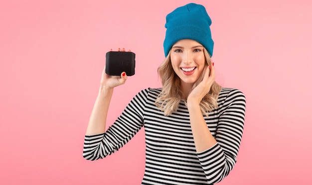 Young woman holding wireless speaker listening to music wearing striped shirt and blue hat smiling posing on pink