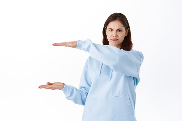 Young woman holding your copy space and grimacing upset, dislike something she holds in hands, showing object and complaining, standing upset against white wall