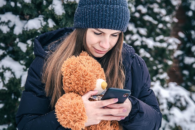 A young woman holds a teddy bear and a smartphone in her hands in snowy weather