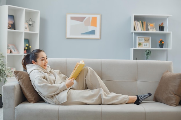 Free photo young woman in home clothes relaxing on a couch at home interior reading a book happy and positive smiling cheerfully