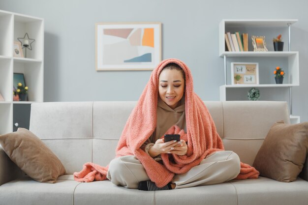 Young woman in home clothes sitting on a couch under blanket texting a message using smartphone looking happy and excited