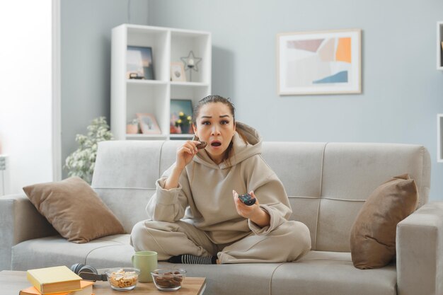 Молодая женщина в домашней одежде сидит на диване в домашнем интерьере и ест печенье, держа в руках пульт дистанционного управления, смотрит телевизор, изумляясь и удивляясь