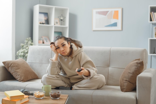 Giovane donna in abiti da casa seduta su un divano a casa interno mangiare cookie tenendo telecomando guardare la televisione annoiata e triste