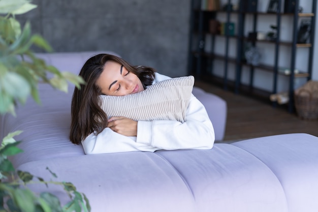 Young woman at home in a white hoodie on the couch calmly hugging a pillow with closed eyes