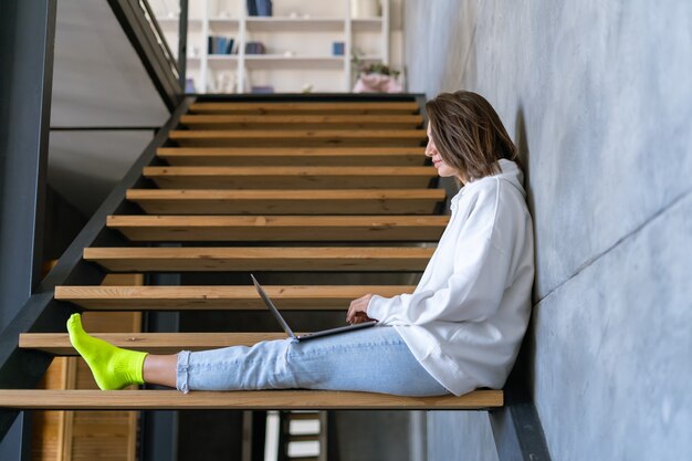 A young woman at home in a white hoodie and jeans sits on the stairs with a laptop on her knees