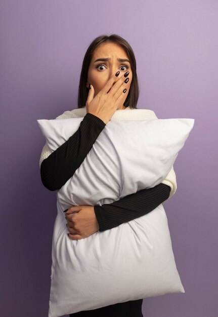 Молодая женщина обнимает подушку в шоке, прикрывая рот рукой