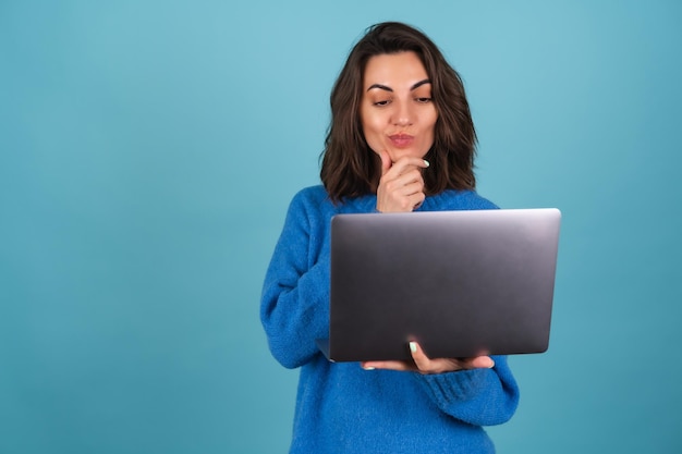 Молодая женщина в вязаном свитере изолирована, держа ноутбук, задумчиво глядя на экран