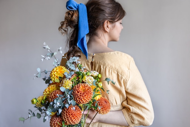 Молодая женщина в желтом платье с голубой лентой в волосах, держа букет желтых и оранжевых хризантем, серый фон.