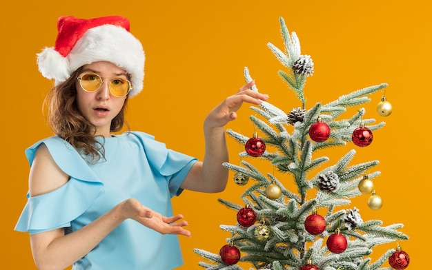 無料写真 青いトップとオレンジ色の背景の上に立って混乱しているように見えるクリスマスツリーを飾る黄色いメガネを身に着けているサンタ帽子の若い女性