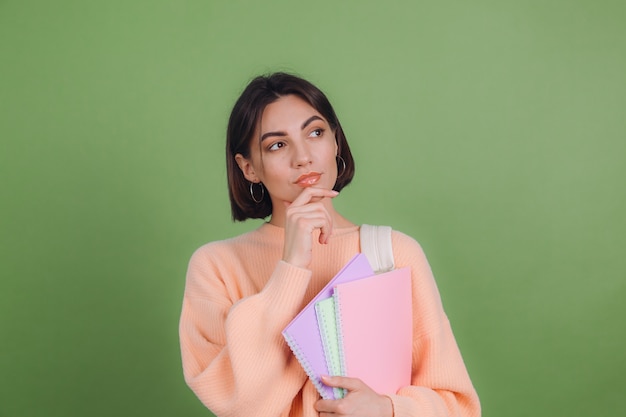 緑のオリーブ色の壁に分離されたカジュアルな桃のセーターの若い女性