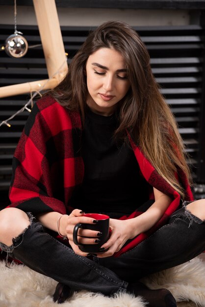 Бесплатное фото Молодая женщина в клетчатом пледе сидит на полу и держит чашку.