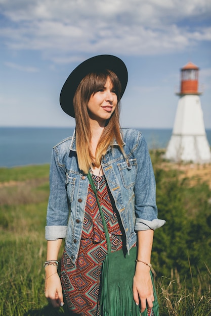 Бесплатное фото Молодая женщина на природе, маяк, богемный наряд, джинсовая куртка, черная шляпа, улыбается, счастливая, лето, стильные аксессуары