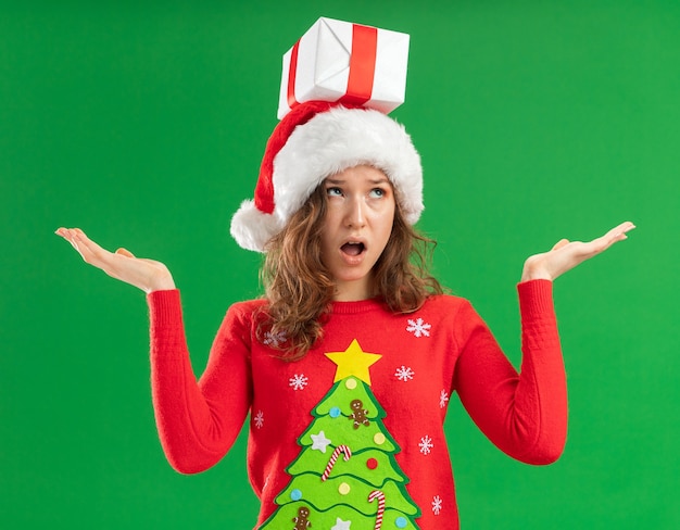 無料写真 赤いクリスマスセーターとサンタの帽子をかぶった若い女性が頭にプレゼントを持って混乱しているように見える緑の背景の上に立っている側に腕を広げて