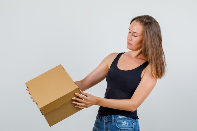 Бесплатное фото Молодая женщина в майке, шортах держит картонную коробку и смотрит внимательно
