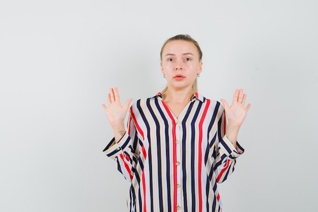 Бесплатное фото Молодая женщина в полосатой блузке показывает жест сдачи, поднимает руки и выглядит испуганной