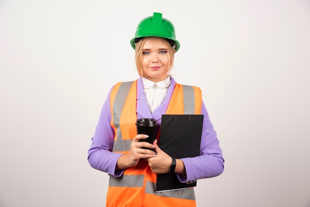 Young woman industrial engineer in uniform with clipboard and black cup on white.