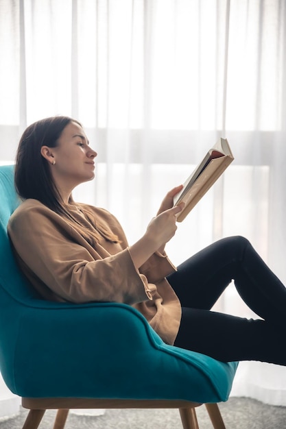 A young woman is reading a book while sitting on an armchair by the window