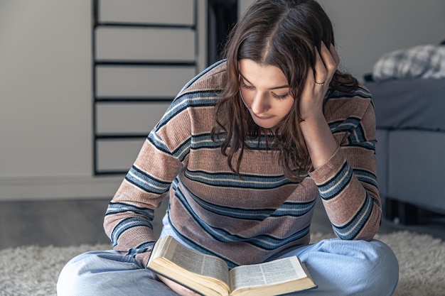 A young woman is reading a book while sitting on the floor in her room