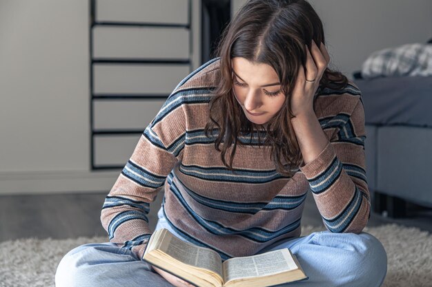Молодая женщина читает книгу, сидя на полу в своей комнате.