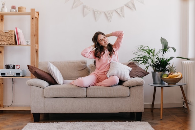 Young woman is resting in her room, sitting on couch, surrounded by bookshelves and table with flower pot