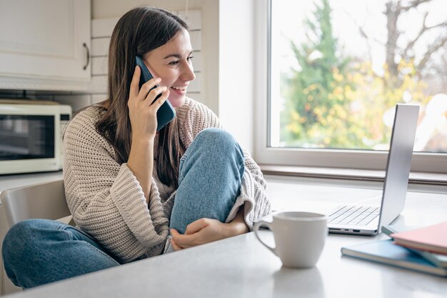 A young woman is sitting in the kitchen with a smartphone and a laptop