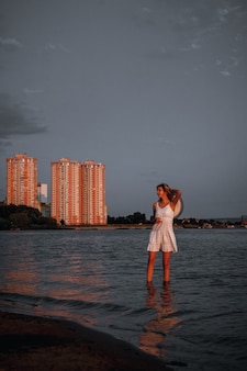 A young woman is standing in the water a beautiful slender happy blonde in a white summer lace dress...