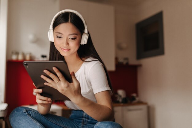 Young woman in jeans and white T-shirt looks into tablet and listens to music in headphones