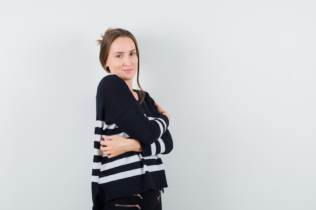 Young woman keeping arms crossed in striped knitwear and black pants and looking happy