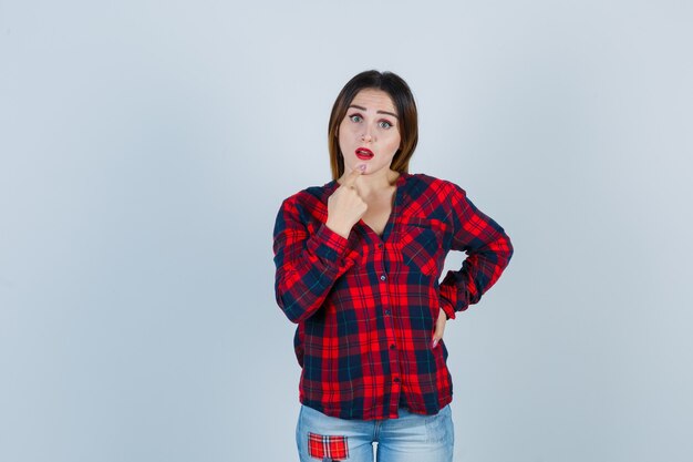 Young woman keeping finger on chin in checked shirt and looking surprised , front view.