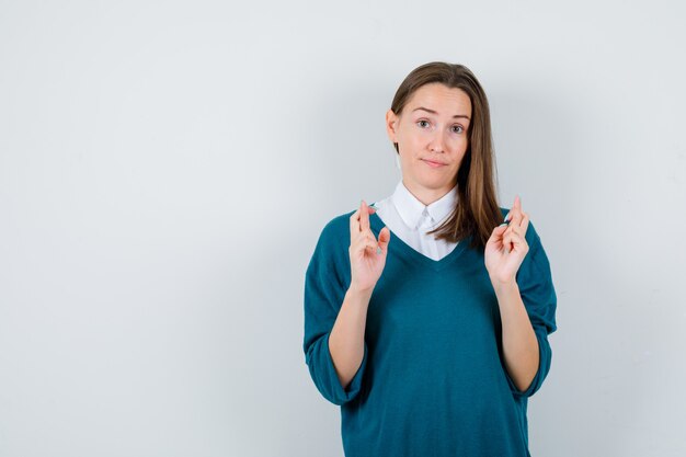 Young woman keeping fingers crossed in sweater over white shirt and looking clueless , front view.