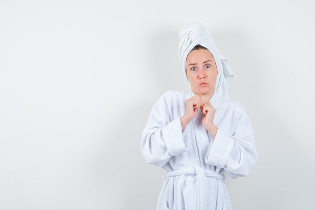 Young woman keeping fists over chest in white bathrobe, towel and looking scared , front view.