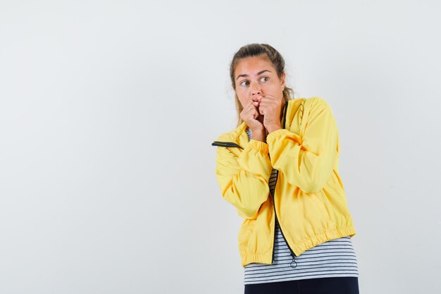 Young woman keeping fists on mouth in t-shirt, jacket and looking scared , front view.