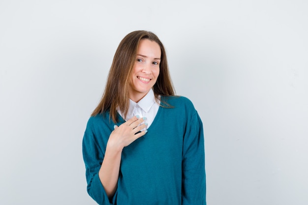 Young woman keeping hand on chest in sweater over white shirt and looking cheerful. front view.