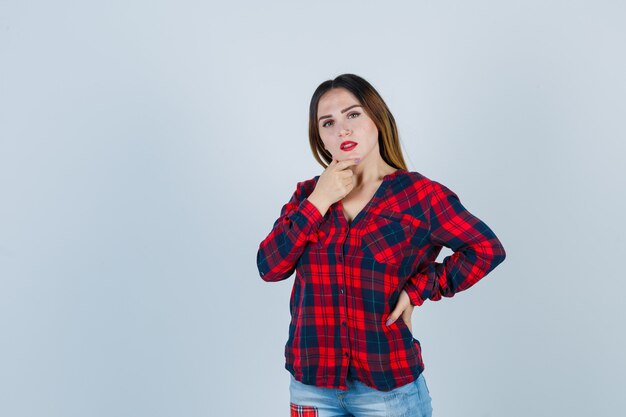 Young woman keeping hand on chin in checked shirt and looking thoughtful , front view.
