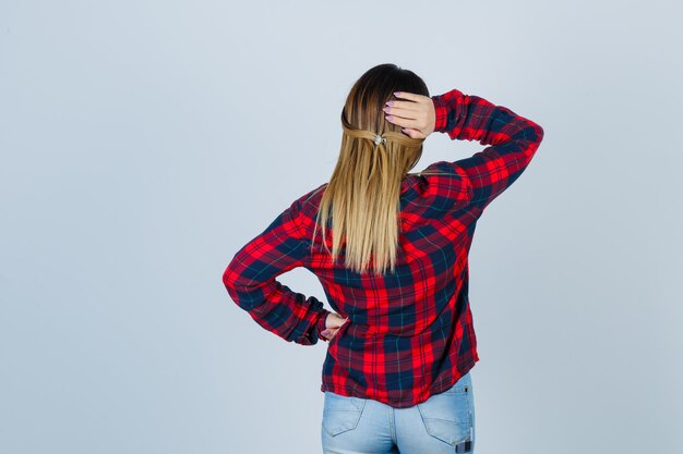 Young woman keeping hand behind head in checked shirt, jeans and looking pensive , back view.