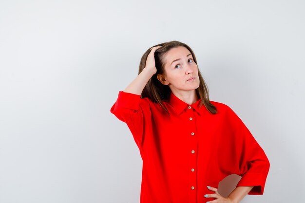 Young woman keeping hand on head, looking up in red blouse and looking thoughtful. front view.