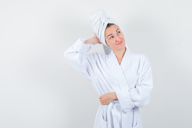 Young woman keeping hand behind head in white bathrobe, towel and looking forgetful. front view.