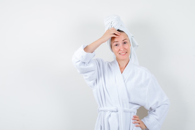 Young woman keeping hand on head in white bathrobe, towel and looking merry. front view.