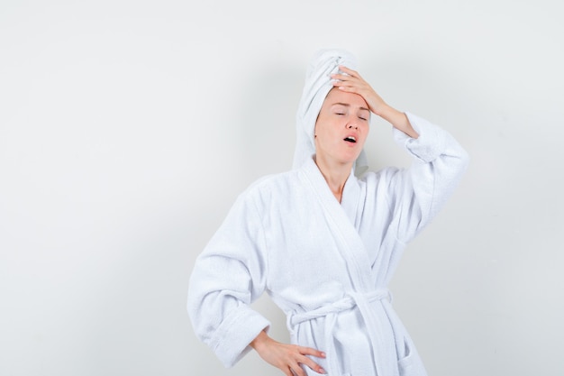 Young woman keeping hand on head in white bathrobe, towel and looking sorry , front view.