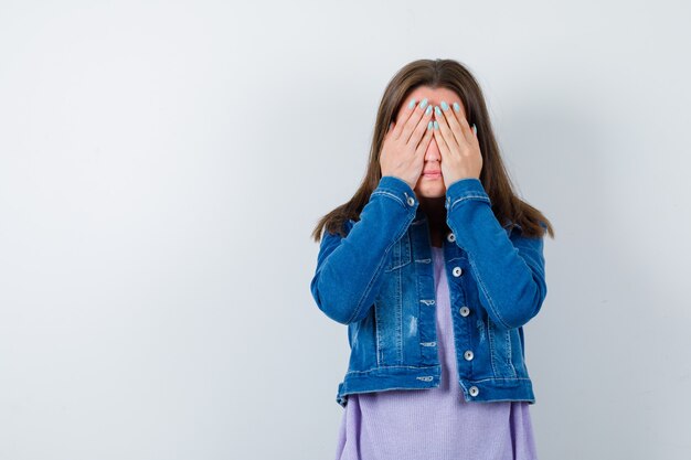 Young woman keeping hands on eyes in t-shirt, jacket and looking scared. front view.