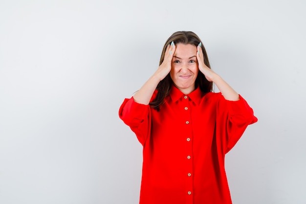 Young woman keeping hands on head in red blouse and looking forgetful. front view.