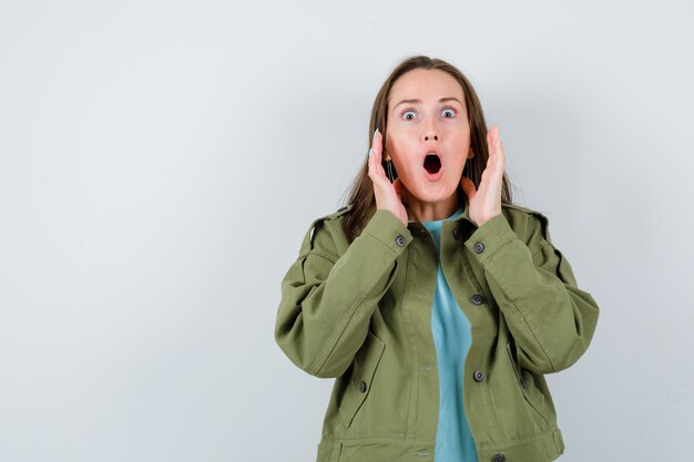 Young woman keeping hands near face in green jacket and looking shocked. front view.