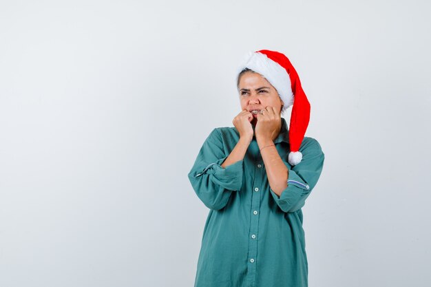 Young woman keeping hands near mouth in shirt, Santa hat and looking terrified . front view.