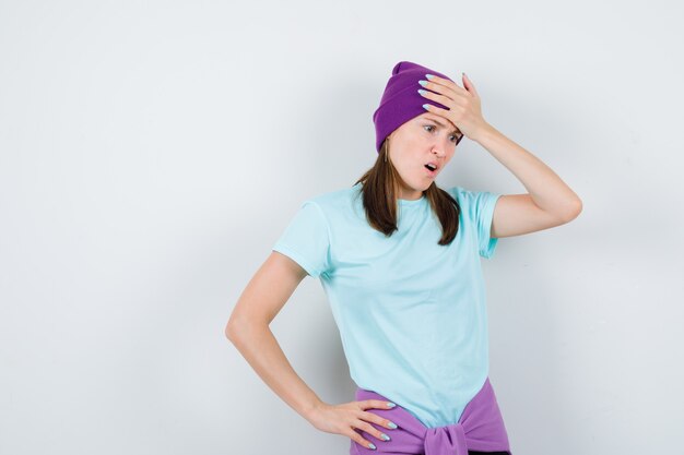 Young woman keeping one hand on forehead, another hand on hip in blue t-shirt, purple beanie and looking shocked. front view.