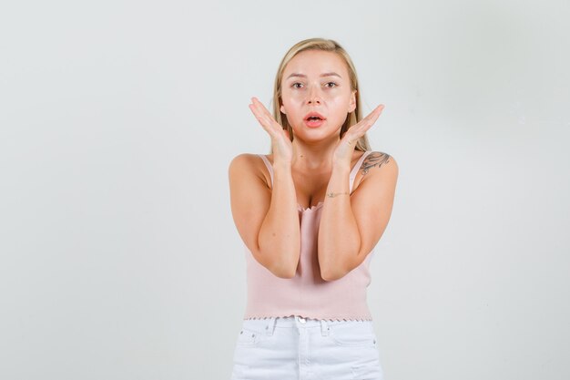 Young woman keeping palms near face in singlet, mini skirt and looking scared 